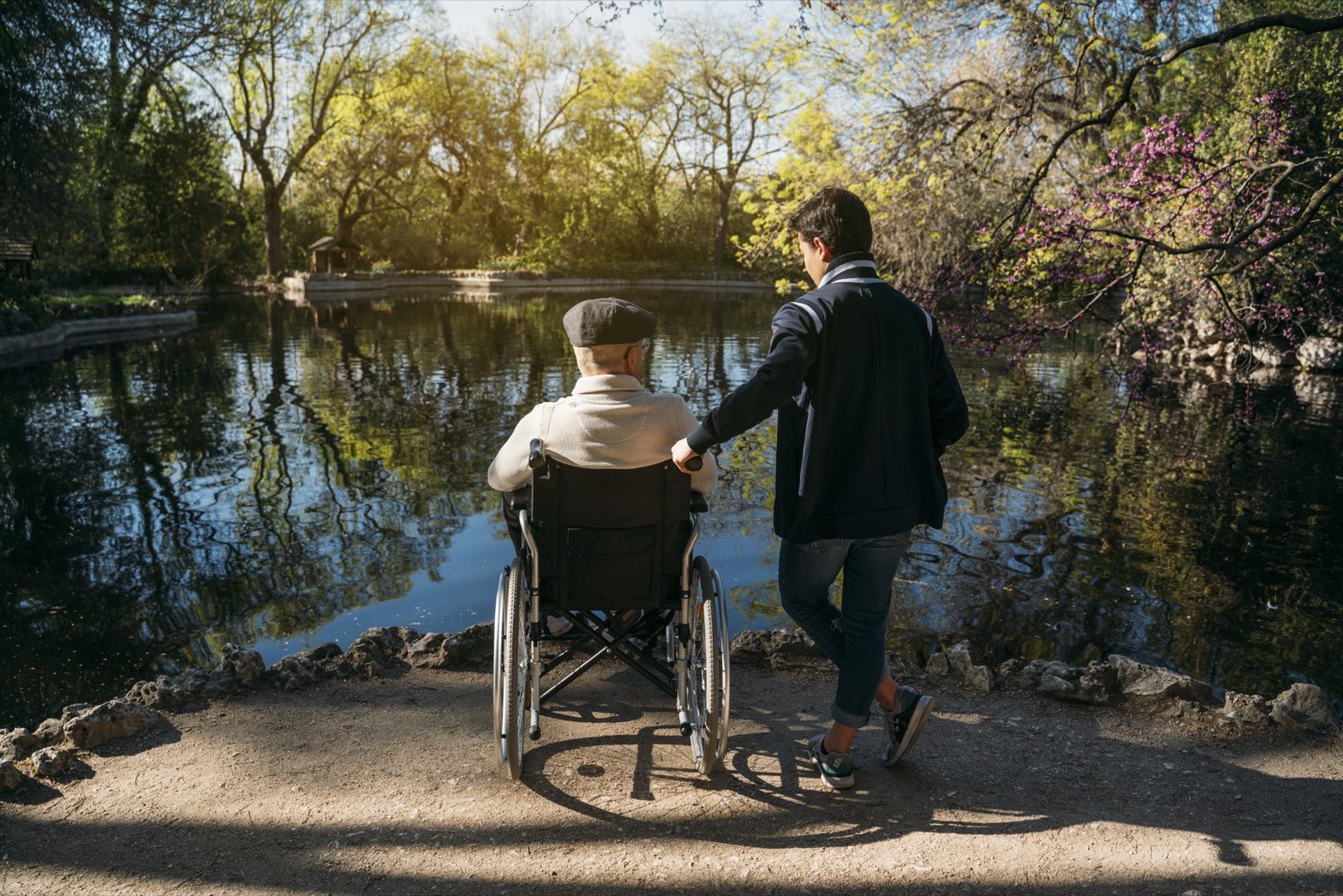 A support worker and an older man in a wheelchair looking out over a pond