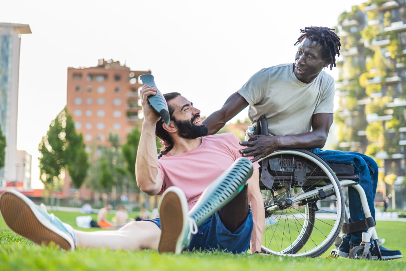 Two friends laughing together on the grass in a park, one using a wheelchair and one with prosthetic legs