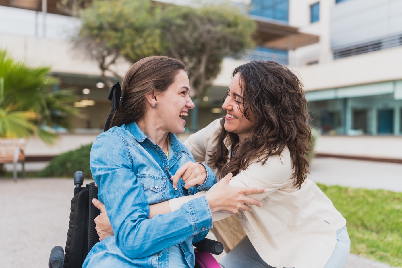 Two women greeting each other warmly outdoors, one using a wheelchair