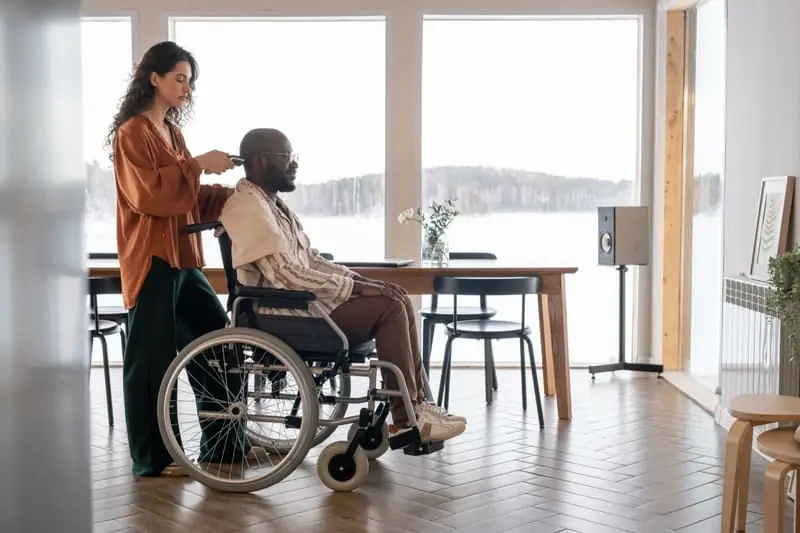 A support worker trimming the hair of a man seated in a wheelchair at home, in front of a bright window